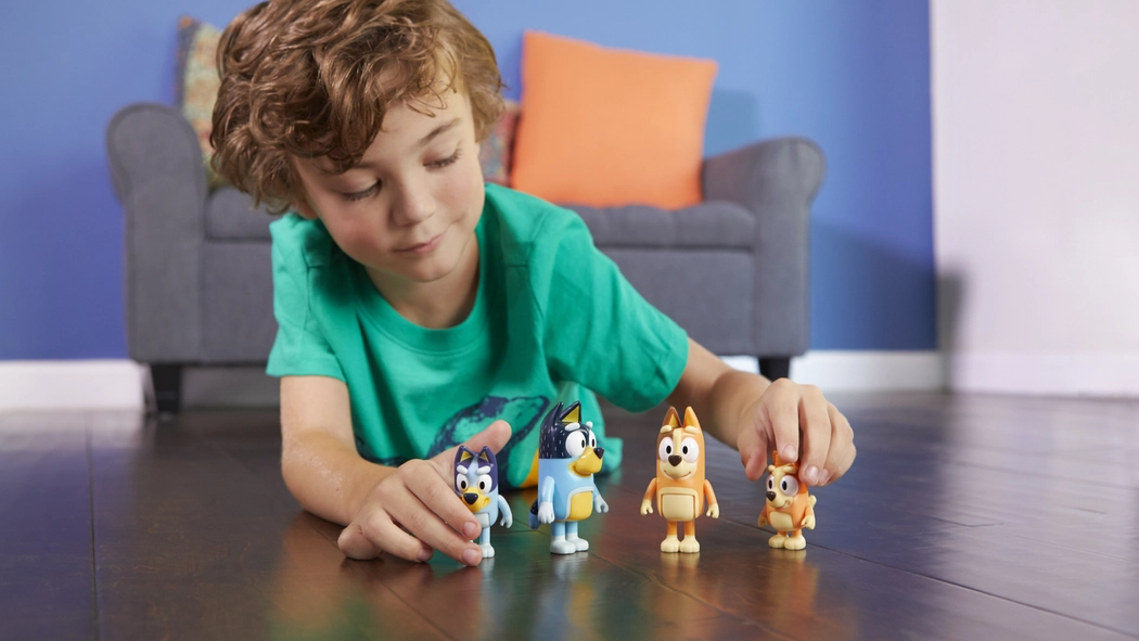 Young boy in green t-shirt lying across the floor whilst playing with four small Bluey family figurines.
